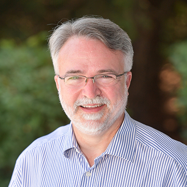 James Venhaus '91 faces the camera in front of outdoor scenery. He is wearing a striped shirt and glasses.