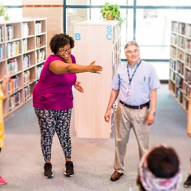 Jasmine Thomas '12 (left) gestures to something out of frame. A man stands to the right. They are in a children's library.