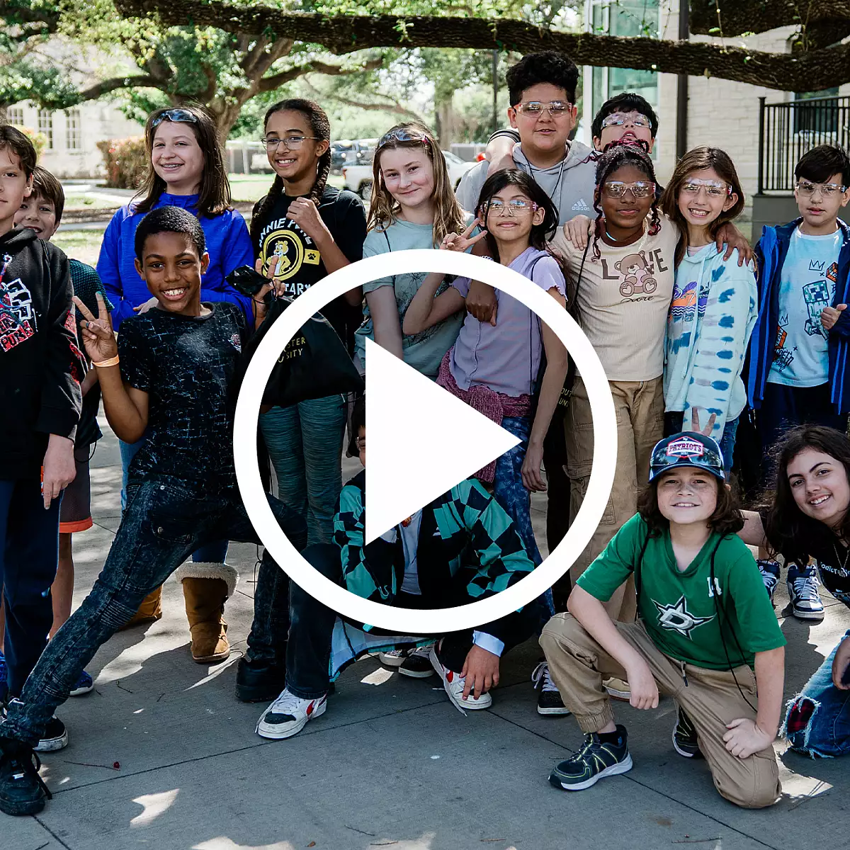 Group of smiling elementary school students posing together outdoors under a tree, overlayed with a large play button icon.